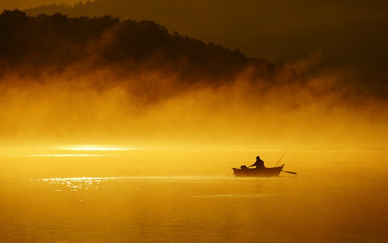 Bei Sonnenaufgang ist ein Angler in seinem Ruderboot mitten auf einem See zu sehen. Der See liegt in goldenem Licht.