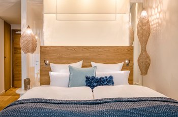 Übernachten in der Seezeitlodge Ein gemütlich beleuchtetes Zimmer mit einem großen Doppelbett mit einer blauen Tagesdecke und vielen Kissen darauf.