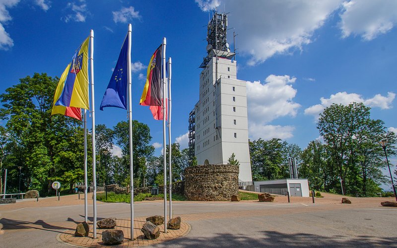 Der weiße Schaumbergturm steht auf dem Schaumberg. Davor befindet sich ein Buswendeplatz mit Fahnen.