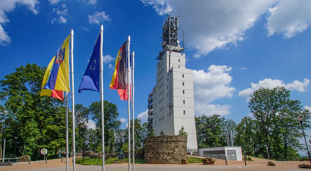 Der weiße Schaumbergturm steht auf dem Schaumberg. Davor befindet sich ein Buswendeplatz mit Fahnen.