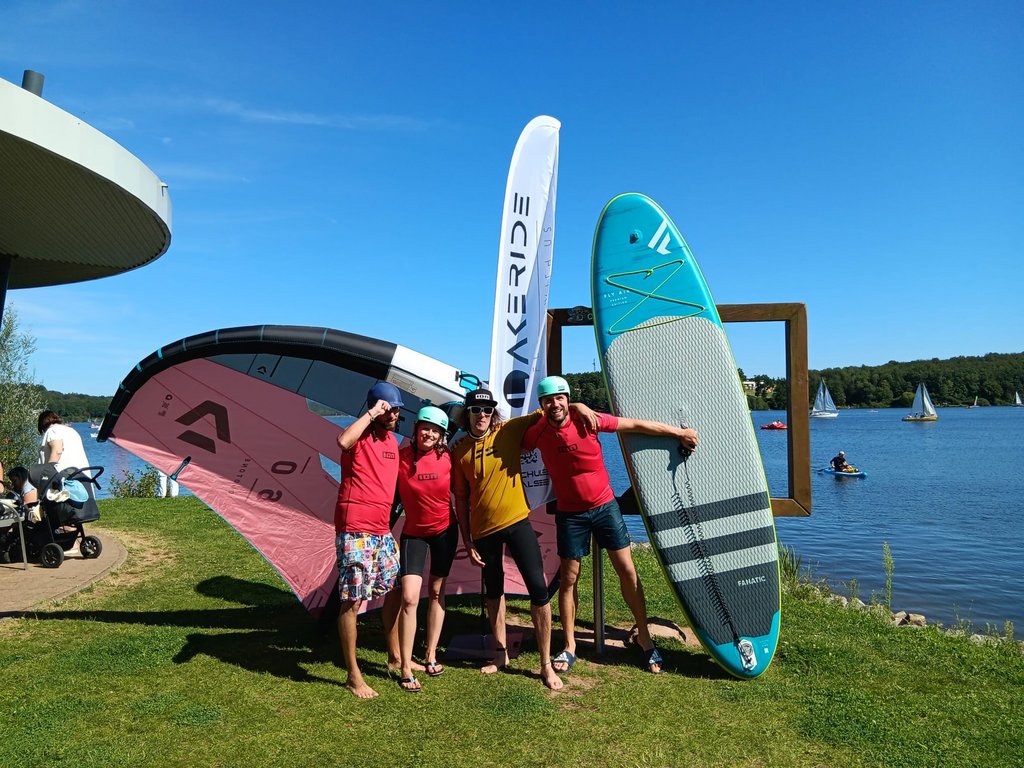 4 Personen stehen am Ufer des Bostalsees und haben ein Wingfoil, ein SUP und eine Beachflag mit dabei.