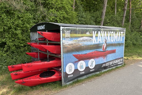 Eine Kajak Verleihstation an einem Weg. In einem überdachten Gehäuse sind mehrere Kajaks sowie SUP-Boards gelagert.