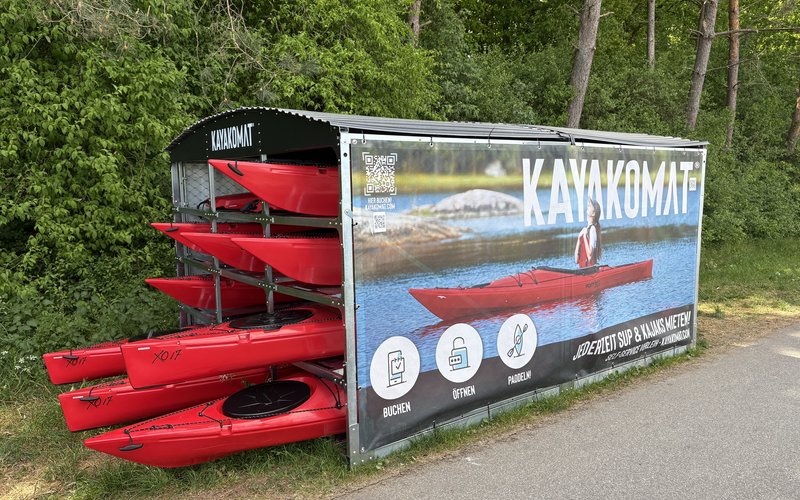Eine Kajak Verleihstation an einem Weg. In einem überdachten Gehäuse sind mehrere Kajaks sowie SUP-Boards gelagert.