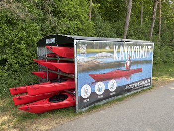 Eine Kajak Verleihstation an einem Weg. In einem überdachten Gehäuse sind mehrere Kajaks sowie SUP-Boards gelagert.