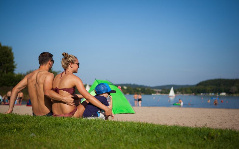 Ein junges Paar mit kleinem Kind sitzt auf der Liegewiese eines Stranbads am Bostalsee und schaut auf den See. Im Hintergrund ist eine grüne Strandmuschel zu sehen.