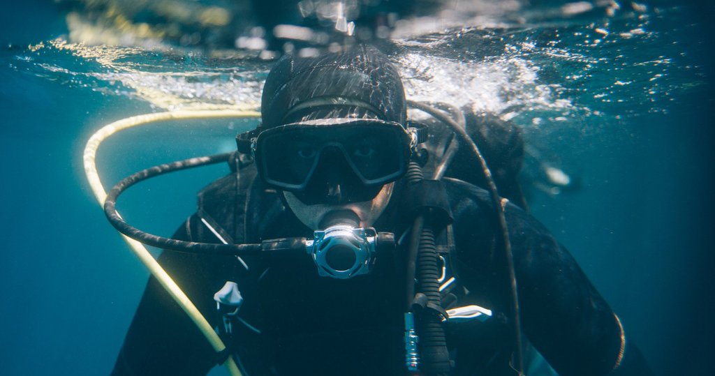 Ein Mensch in Taucheranzug und Taucherbrille und Sauerstoffmaske befindet sich unter Wasser. 
