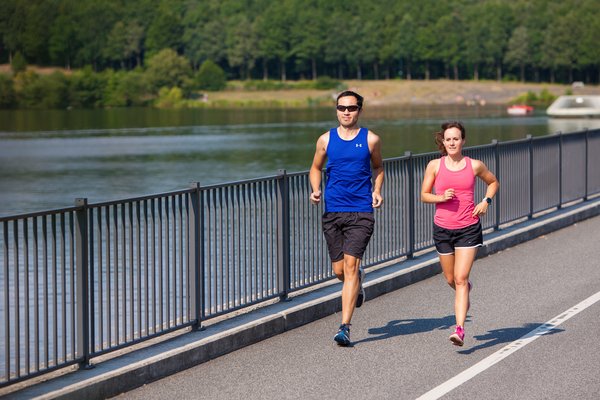 Eine Frau und ein Mann in Sportkleidung joggen auf dem Staudamm des Bostalsees. Hinter ihnen breitet sich das Seeufer und davor das Wasser des Bostalsees aus.