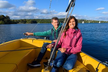 Eine Frau und ein Herr in Outdoorkleidung sitzen auf dem geleben Segelboot der Segelschule S.A.L.T. Yacht GmbH. Die Frau zieht die Segel an, während der Herr das Boot am Heckruder steuert. Beide schauen zufrieden in Richtung Kamera. In der Ferne sind weitere Segelboote auf dem Wasser zu sehen.