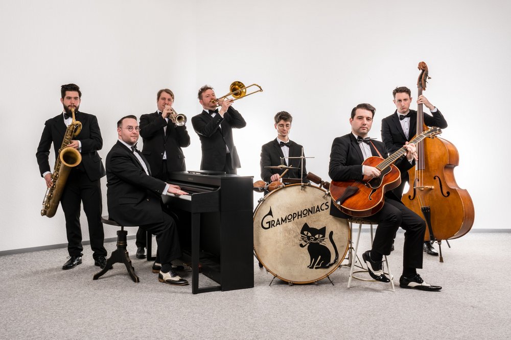 Eine Band, bestehend aus 7 Männern im Anzug, spielt auf verschiedenen Musikinstrumenten.