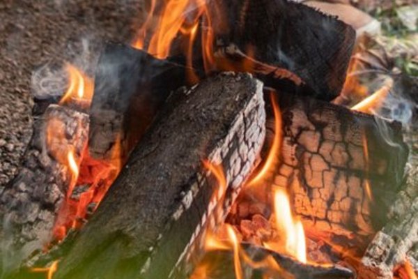 Vor einem Mietwohnwagen des Campingplatzes Bostalsee hat es sich ein Pärchen mit einem Getränk am Grillfeuer, über dem ein Schwenker hängt, gemütlich gemacht.