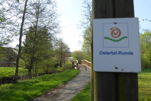Ein asphaltierter Weg führt über eine Brücke zu einem Gebäude. An einem Pfosten vorne rechts hängt ein Wegweiser mit der Aufschrift "Ostertal-Runde".